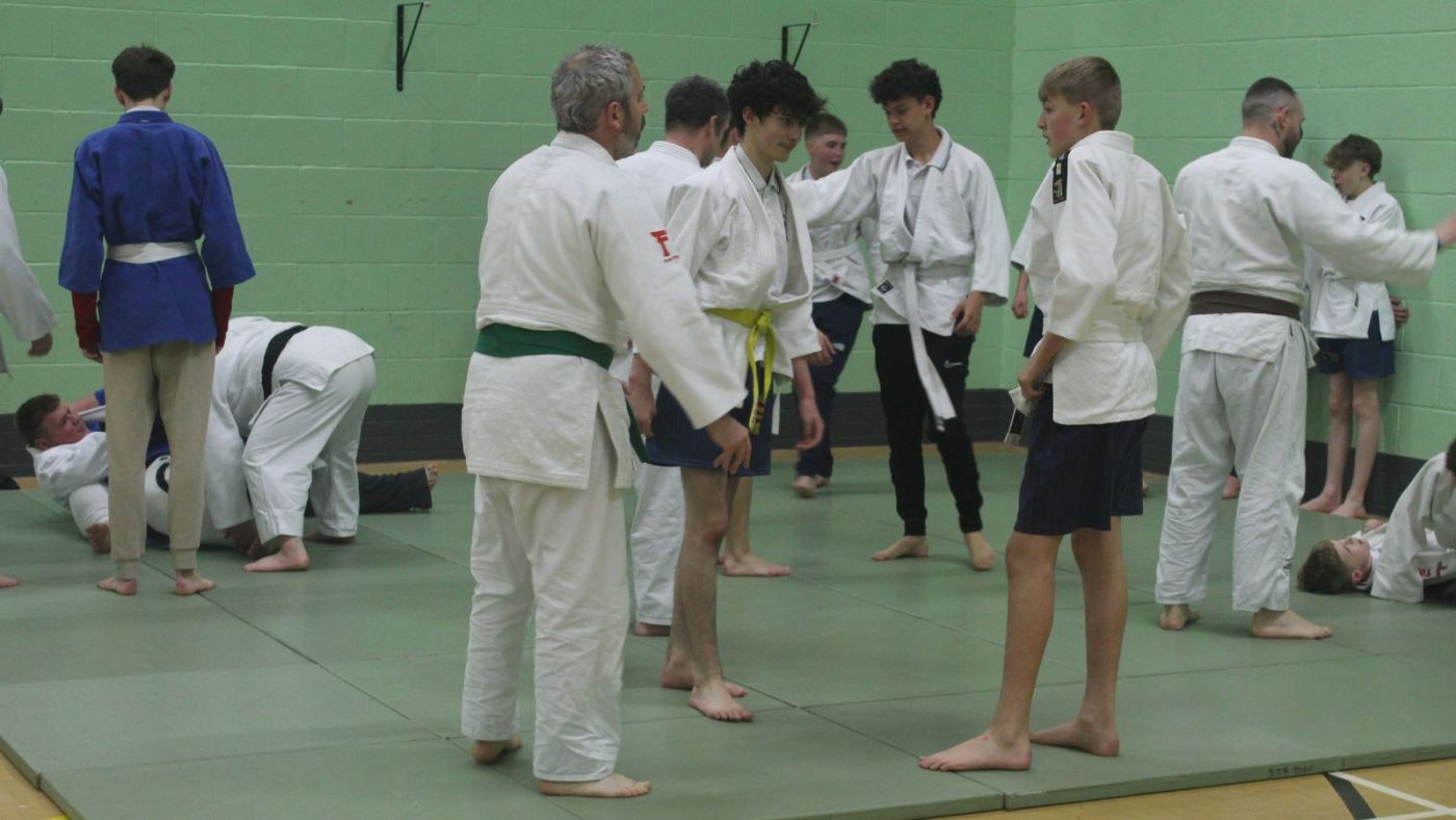 Secondary school pupils taking part in judo. Two boys listening to teacher explain with other pupils stood around them in sports hall.